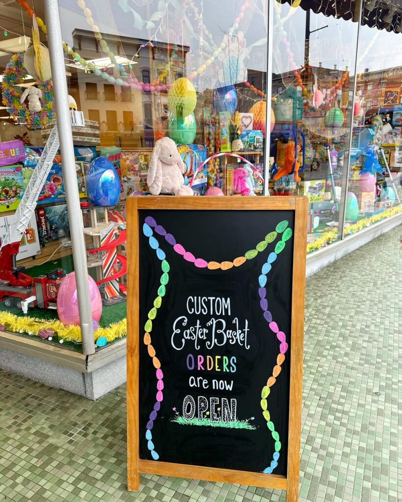Chalkboard sign that says "Custom Easter Basket Orders are Now Open" in front of one of our store's Easter Display Windows